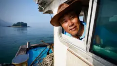 Um homem usando chapéu aparece com a cabeça para fora da janela de um barco. Ao fundo, vê-se o mar e uma ilha.