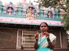 Shilpa está em pé diante de um templo com deuses hindus decorativos, segurando seu colar de contas e pérolas