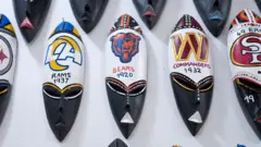 Uma fileira de máscaras tradicionais ganesas com os logotipos e nomes de times de futebol americano — Rams, Bears e Commanders — pintados por cima.