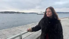 Una mujer con un abrigo marrón mira hacia el agua en un día frío y gris en Oslo.