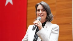 Sinem Dedetaş elinde mikrofonla sahnede konuşuyor. Beyaz bir takım giymiş, arkasında Türk bayrağı var.