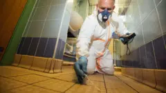 Ben Giles fotografiado arrodillado en un baño con azulejos azules. Lleva equipo de protección personal blanco completo, guantes de látex azules y sostiene un hisopo de algodón contra las baldosas del suelo. En la otra mano sostiene un dispositivo negro. Lleva una mascarilla y tiene el pelo corto y canoso. Al fondo, se ven dos lavabos blancos.