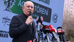 Erkol Ankara'da katıldığı bir programda kürsüde konuşuyor. Üzerinde siyah takım elbise, elinde mikrofon var.