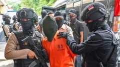 Seorang terduga teroris dengan wajah tertutup sedang dibawa polisi bersenjata.