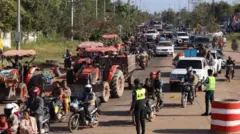 乗用車やトラクター、トラック、バイクなどで渋滞している道路。道のわきは緑が生い茂っている。蛍光色のベストを着て交通整理をしている人もいる