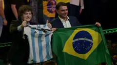 Javier Milei segura uma camisa da seleção argentina durante evento em São Paulo que oficializou Flávio Bolsonaro como candidato. Flávio está ao lado de Milei segurando uma bandeira do Brasil.