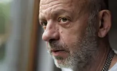 Antony Easton, um homem careca com barba grisalha. Seu rosto está levemente virado para o lado, destacando rugas. Ele veste uma camisa branca e um colar de corrente prateada. O fundo está suavemente desfocado.