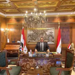 صبري نخنوخ يجلس على كرسي وأمامه مكتب خشبي مزكرش بلون ذهبي وحوله أعلام مصر وخلفه لوحة، ويبدو أن أثاث المكتب ثمين. وفي السقف إضاءة كريستالية كبيرة. يرتدي بدلة رسمية تميل إلى اللون البني مع قميص أبيض وربطة عنق بنفس لون البدلة منطقة كما يبدو باللون بالذهبي.