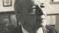 A imagem em preto e branco mostra um retrato em close de um homem idoso, Mario Quintana, de cabeça parcialmente calva, usando terno e camisa social. Ele aparece com o olhar voltado para cima e para a direita, em expressão serena e reflexiva, enquanto o fundo desfocado revela quadros pendurados na parede e outra pessoa sentada ao longe.