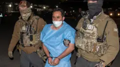 Homem em roupa prisional azul, Mohammad Sharifullah, com máscara cirúrgica e algemado, sendo levado à noite por pelos braços dois homens vestindo uniformes táticos do FBI, com um avião ao fundo