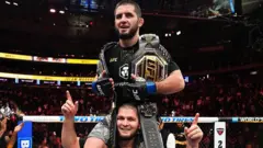 اسلام ماخاچف با پیراهن سیاه درحالی که کمربند قهرمانی در دست دارد، روی شانه‌های حبیب نورمحمدوف مربی‌اش قرار دارد