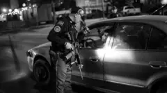 Um policial armado com uma lanterna no rosto de uma pessoa que está dentro de um carro