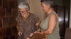 Mujer ayuda a caminar a anciana en una casa
