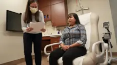 Una enfermera coordinadora de trasplantes de corazón junto a Jessica López, sentada en una silla en la habitación del hospital.