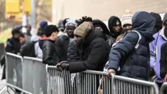 Large number of pipo wey wear black hoodies, one man put hand on di iron barricade wit head warmer.