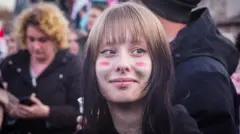 Burnu delik bir genç kız, kahkülleri var. Yanaklarına macar bayrağı renkleri boyalı