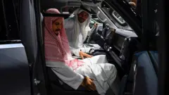 在一場車展上，兩名身穿長袍、頭戴頭巾的男子，分別坐在一輛中國製造的汽車的駕駛座位和前排乘客座位。