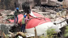 Recovery workers stand beside the tail section of the Air India 787 plane which is in situ at the crash site in Ahmedabad. They are winching the fuselage which is painted white and red - the Air India colours. The tail fin has detached and other debris surrounds the fuselage