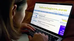 Woman using a laptop while viewing the FabSwingers website. The screen shows forum and message board pages. The image is taken over the woman's shoulder in a dimly lit indoor setting.