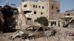 Imagem de um carro totalmente destruído no meio de escombros. No fundo, um prédio igualmente destruído.