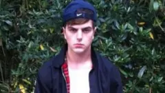 Will Adolphy na adolescência. Ele veste uma camiseta branca, uma jaqueta azul com zíper e um boné azul-escuro virado para trás. Olha com raiva para a câmera.