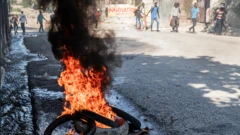 Cadáver arde sob pneus em uma rua do centro de Porto Príncipe, no dia 9 de fevereiro de 2026, com pessoas desfocadas ao fundo