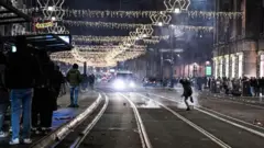 Okićene ulice, ljudi na trotoarima, dim od vatrometa po šinama 