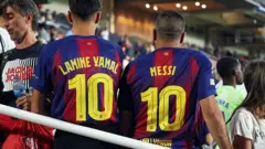 Dois torcedores do Barcelona vestem camisas de futebol com o número 10 nas costas: à esquerda, a camisa leva o nome de Lamine Yamal; à direita, o de Lionel Messi.