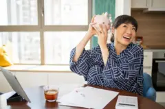 Mulher segurando cofrinho, sorrindo e com papéis e calculadorasimulador de casa de apostascima da mesa