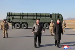Kim Jong-un, flanqueado por una mujer joven y seguido por varios militares norcoreanos, camina alejándose de un vehículo militar pesado.