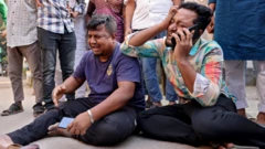 রাস্তায় বসে পা ছড়িয়ে কাঁদছেন দুই জন, তাদের একজন কানে মোবাইল ফোন ধরে আছেন। পেছনে কয়েকজন মানুষ তাদের ঘিরে দাঁড়িয়ে আছেন