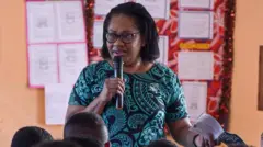 Uma mulher de vestido preto e turquesa, usando óculos e falando ao microfone diante de várias pessoas. Atrás dela, uma parede com papéis colados