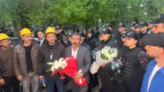 Elinde emniyet görevlilerinden aldığı çiçeği tutan biri, yanında sarı kask takan madenciler ve polisler var.