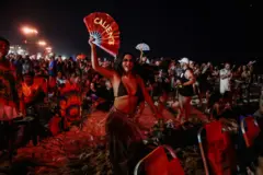 Mulher segura leque com a palavra caliente. Ela está na praia, à noite. Muita gente atrás.