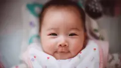 A baby stares at the camera, smiling from her cot. She wears a pink bib with flower motifs and appears calm, but intent