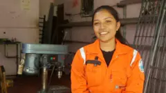 වෙල්ඩින් කරන ස්ථානයක තැඹිලි පාට ඇඳුමක් ඇඳගත් තරුණියක් සිනාමුහු මුහුණින් සිටී.