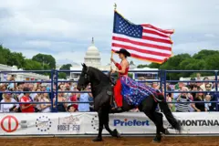 elinde amerikan bayrağı tutan bir kadın at üzerinde renkli rodeo kıyafetiyle, etrafında insanlar onu izliyor ve arkada kongre binası görülüyor