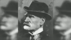 Foto en blanco y negro de Cobb Morley, de perfil, con un sombrero, camisa de cuello alto, corbata antigua y chaqueta, estilo victoriano. lleva barba y bigote.