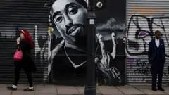 Uma mulher caminha enquanto outra pessoa está diante de um mural do astro do hip-hop Tupac Shakur, em Londres, em 2024