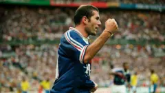A imagem mostra um jogador da seleção francesa comemorando de forma vibrante durante uma partida, com o punho cerrado e expressão de entusiasmo. Ao fundo, um estádio lotado aparece desfocado, enquanto jogadores brasileiros podem ser vistos à distância, reforçando o contexto de um confronto internacional.