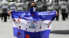Uma mulher segura uma bandeira da Nicarágua com manchas vermelhas simbolizando sangue, durante um protesto contra o governo em 2018. Ela usa um gorro preto, óculos escuros tipo aviador e tem parte do rosto coberto por um pano nas cores da bandeira da Nicarágua