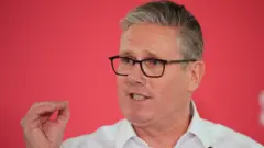 Primeiro-ministro britânico usando óculos com uma armação preta e uma camisa branca. Ele gesticula enquanto fala