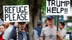 Dois manifestantes segurando cartazes em que se lê: "REFÚGIO, POR FAVOR" e "AJUDA, TRUMP!!" do lado de fora da embaixada americana em Pretória, na África do Sul, em fevereiro