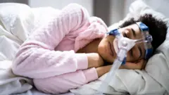 Una mujer joven duerme de lado en la cama con una máscara que cubre su nariz y está conectada a un largo tubo plástico