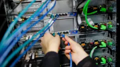 На заднем плане серверы и множество проводов. На переднем плане две руки подключают синий кабель к серверу.