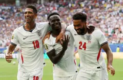 Bukayo Saka akoze ikimenyetso kigaragaza intoki eshatu, yishimana na bagenzi be b'ikipe y'igihugu y'Ubwongereza, ubwo yari amaze kuyitsindira igitego cya gatanu, cya gatatu cye muri uwo mukino, aha ari kumwe na Jude Bellingham na Reece James, mu mukino wo guhatanira umwanya wa gatatu mu gikombe cy'isi cya 2026 Ubwongereza bwatinzemo Ubufaransa ibitego 6-4, wabereye ku kibuga cya Miami, muri leta ya Florida muri Amerika, ku itariki ya 18 Nyakanga (7) mu mwaka wa 2026.