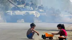 جاده خاکی با ردیف تانکها و دو کودک در کنار جاده در حال بازی با یک کامیون اسباببازی