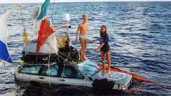 Dos hombres —Marco Amoretti y Marco De Candia— están de pie sobre el capó de un coche que flota en el océano. Al coche le cuelgan redes rojas, cuerdas y banderas. Llevan las piernas al descubierto, y uno de ellos está desnudo, salvo por unos calzoncillos blancos. El otro lleva una camiseta azul marino. Tienen barba y pelo largos, y son delgados y morenos.