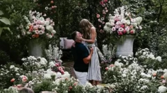 Em um jardim frondoso com flores brancas e rosas, um homem vestido de calças brancas e uma camisa escura de manga curta se ajoelha em frente a uma mulher loira, com um vestido listrado branco e preto sem mangas, que toma seu rosto com as mãos