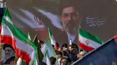 A large photo of Iran's new Supreme Leader, Ayatollah Mojtaba Khamenei, the son of the late Ayatollah Ali Khamenei, is displayed against a national flag and an Islamic Revolutionary Guard Corps (IRGC) flag. Supporters can be seen waving the flags